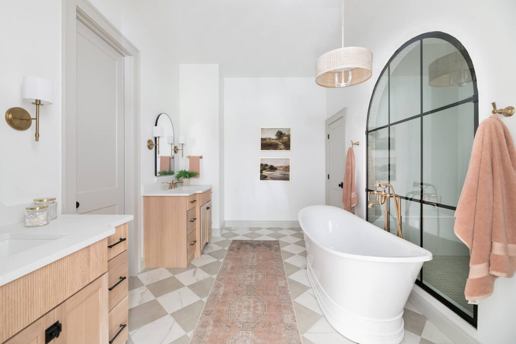 2023 St. Jude Dream Home master bathroom with a large bathtub and dual vanities with the show room visible through the window behind the tub. Sconces sit on the wall framing the vanities and a hanging figure sits above the bathtub.